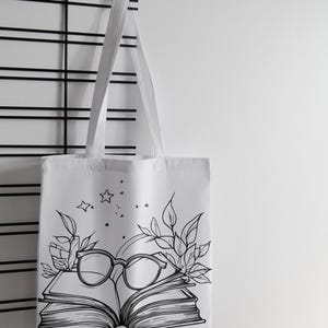 May include: White canvas tote bag with a black and white illustration of a book, glasses, and leaves. The illustration includes stars.