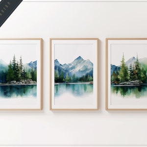 May include: Three framed watercolour paintings depicting a mountain lake scene with trees and mountains reflected in the water. The paintings are in wooden frames and are hung on a white wall.
