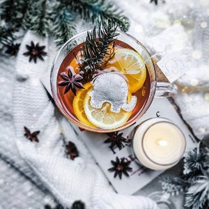 Peut inclure: Une tasse en verre remplie de thé, de tranches de citron et d'anis étoilé. Un sachet de thé en forme d'éléphant blanc est visible dans la tasse. La tasse est posée sur une couverture en tricot blanc avec un livre et une bougie allumée.