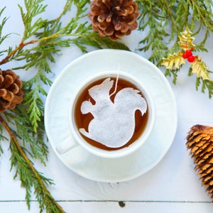 Könnte beinhalten: Eine weiße Teetasse mit Untertasse steht auf einer weißen Oberfläche. Die Teetasse enthält braunen Tee mit einem weißen Teebeutel in Form eines Eichhörnchens. Die Teetasse ist von grünen Kiefernästen und Tannenzapfen umgeben.