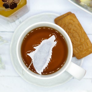 Peut inclure: Une tasse à thé en céramique blanche avec un sachet de thé en forme de loup dans une tasse de thé brun. La tasse à thé est sur une soucoupe blanche.