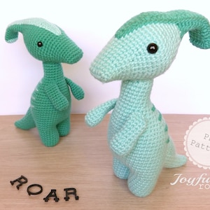 May include: Two crocheted green dinosaur toys with black eyes. The word "ROAR" is spelled out in black letters on a wooden surface. The text "PDF Pattern Joyful round" is on a white circle with a pink border.