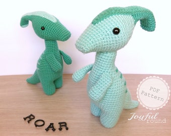 May include: Two crocheted green dinosaur toys with black eyes. The word "ROAR" is spelled out in black letters on a wooden surface. The text "PDF Pattern Joyful round" is on a white circle with a pink border.