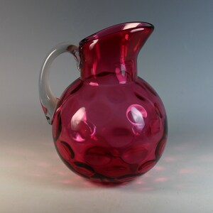 May include: A ruby red glass pitcher with a clear glass handle. The pitcher has a round body with a textured surface of small, raised circles.