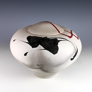 May include: A rounded ceramic vessel with a matte off-white finish, featuring a circular lid. The surface is decorated with a bold, abstract black ink-like splash pattern and a distinct red geometric line design that extends across the lid.