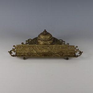 May include: An ornate brass inkwell with a lid and a long, rectangular pen rest. The inkwell and pen rest are both decorated with intricate floral patterns.
