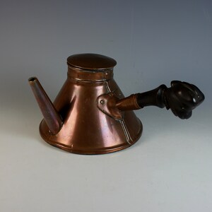 May include: A copper coffee pot with a wooden handle. The pot has a spout and a lid. The handle is shaped like a hand.