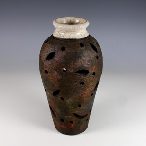 May include: A handcrafted ceramic vase with a dark brown, textured body and a cream-colored, cracked rim. The vase features leaf-shaped cutouts and circular holes, adding a unique decorative element. The vase has a metallic sheen.