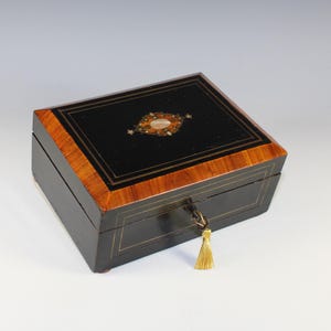 May include: A rectangular black wooden box with a decorative inlaid design on the lid. The box has a key with a gold tassel. The edges of the box are trimmed with a lighter wood and gold accents.