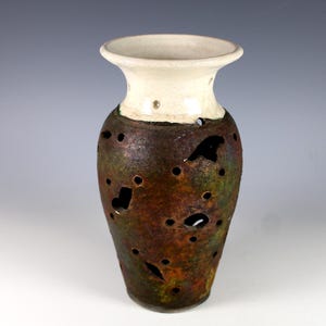 May include: A handcrafted ceramic vase with a cream-colored rim and a textured, dark brown body. The vase features numerous irregular holes and a gradient of green and bronze hues, creating a unique, artistic appearance.