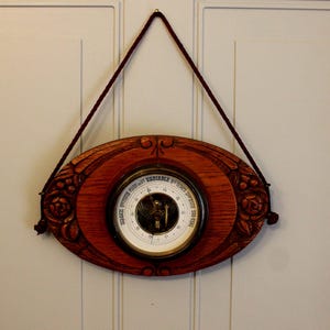 May include: An antique wooden barometer with an oval frame and carved floral details. The barometer face is white with black text and a central needle. A dark red cord is used for hanging.
