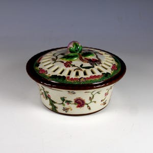 May include: A ceramic container with a lid, featuring a floral and bird design. The container is white with brown trim and a green and red floral border. The lid has a pierced design and a decorative knob.