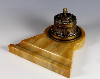 May include: A brass inkwell with an ornate design sits on a yellow marble base. The inkwell has a domed lid with a finial.