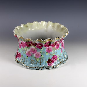 May include: A porcelain bowl with a ruffled, scalloped rim featuring a light blue background decorated with pink and magenta floral patterns, green leaves, and gold filigree accents along the edges and base of the vessel.