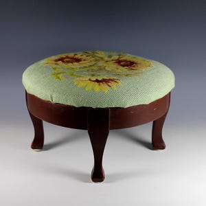 May include: A round footstool with a dark brown wooden frame and four curved legs. The top is upholstered in a light green fabric featuring a needlepoint floral pattern with yellow and brown sunflowers, providing a textured surface.