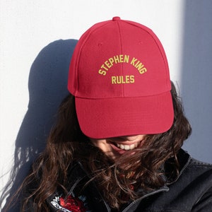 May include: A red baseball cap with the text "Stephen King Rules" embroidered in yellow.