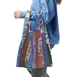 May include: A patchwork tote bag with a variety of patterns and colors, including floral, polka dots, and abstract designs. The bag is held by a person wearing a blue hoodie.