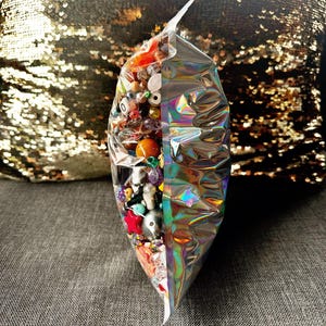 May include: A clear, iridescent bag filled with a variety of colorful beads. The beads include red, orange, purple, and white, with some having unique shapes and textures. The bag is sealed and sits on a gray surface.