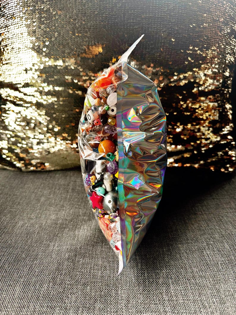 May include: A clear, iridescent bag filled with a variety of colorful beads. The beads include red, orange, purple, and white, with some having unique shapes and textures. The bag is sealed and sits on a gray surface.
