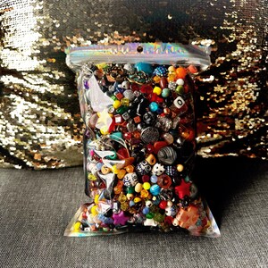May include: A clear plastic bag filled with a colorful assortment of beads, buttons, and small decorative items. The contents include various shapes, sizes, and colors, including red, blue, yellow, and green. The bag is sealed at the top.