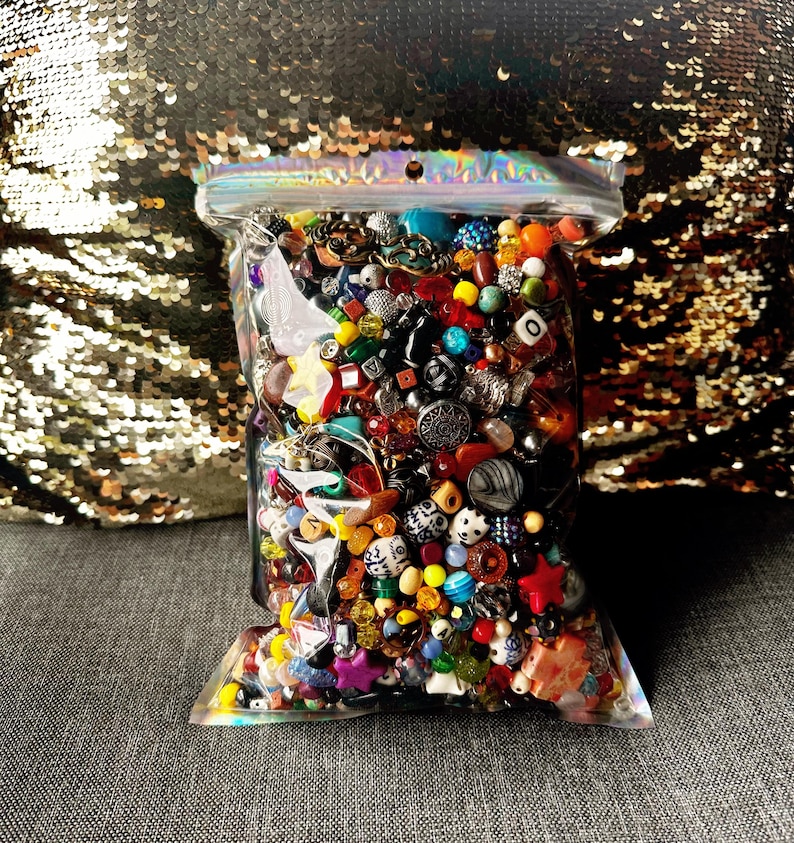May include: A clear plastic bag filled with a colorful assortment of beads, buttons, and small decorative items. The contents include various shapes, sizes, and colors, including red, blue, yellow, and green. The bag is sealed at the top.