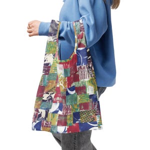 May include: A reusable tote bag with a patchwork design. The bag features a variety of colors including red, blue, green, and yellow. The bag is being held by a person wearing a blue hoodie.