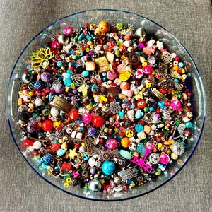 May include: A clear glass serving dish filled with a vibrant assortment of beads, charms, and embellishments in various colors and shapes. Includes red, pink, blue, and yellow beads, along with charms featuring butterflies, stars, and text that reads "Never look back."