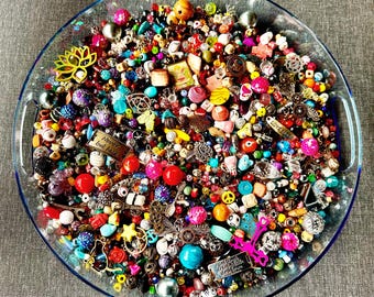 May include: A clear glass dish overflowing with a vibrant assortment of beads, charms, and embellishments in various shapes, sizes, and colors. The collection includes red, pink, blue, and yellow beads, along with metal charms and decorative elements. Some charms have text that reads "Never look back".