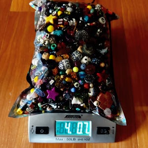 May include: A clear plastic bag filled with a variety of colorful beads, including round, star, and novelty shapes. The bag is on a digital scale, displaying a weight of 4.8 oz. The beads are in shades of red, yellow, blue, and white.