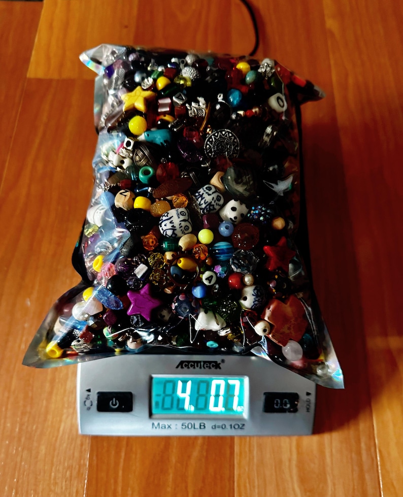 May include: A clear plastic bag filled with a variety of colorful beads, including round, star, and novelty shapes. The bag is on a digital scale, displaying a weight of 4.8 oz. The beads are in shades of red, yellow, blue, and white.