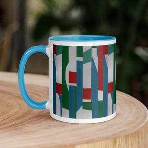 May include: White ceramic mug with a blue handle and interior. The exterior features a colorful geometric pattern with red, green, and blue vertical shapes. The mug is sitting on a wooden surface.