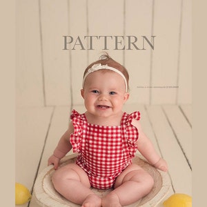 May include: A baby girl wearing a red and white gingham romper with white ruffle trim sits in a white wooden bowl. The baby is smiling and has a white headband with a flower on top.