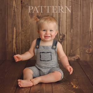 May include: A young child wearing a gray and white striped overall with black straps sits on a wooden floor. The child is smiling and looking at the camera. The background is a wooden wall with the word "PATTERN" written on it.