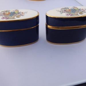 May include: Two navy blue oval-shaped trinket boxes with gold trim. Each box has a white lid decorated with a colorful fruit basket design. The boxes are side by side, showcasing their elegant design and craftsmanship.
