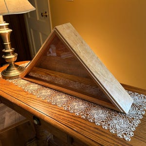 May include: A wooden shadow box with a glass front, designed to hold a folded American flag. The box is sitting on a wooden table with a white lace doily.