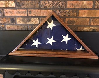 May include: A wooden flag display case with a glass front, showcasing a folded American flag. The flag features white stars on a blue field and red and white stripes. The case is made of dark wood and is triangular in shape.