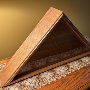 May include: A wooden flag display case in a triangular shape. The case is made of light brown wood with a glass front. The case is designed to hold a folded flag and is displayed on a wooden table.