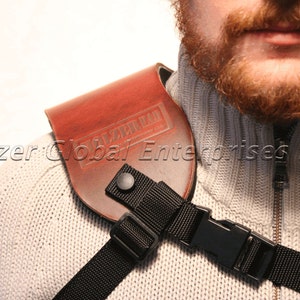 May include: A brown leather shoulder holster with the text "HOLZER GLOBAL ENTERPRISES LLC" embossed on the front. The holster is secured with a black nylon strap and buckle. The person is wearing a gray sweater.