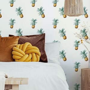 May include: A bedroom with a white wall featuring a pineapple pattern wallpaper. The bed has white bedding, brown and yellow pillows, and a yellow knotted pillow. A wooden side table with a brown glass vase and a yellow vase with dried flowers sits next to the bed.