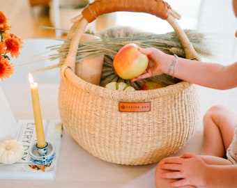 Puede incluir: Una cesta tejida con asa de cuero, llena de manzanas y otras frutas. La cesta tiene una etiqueta de cuero marrón con el texto "Design Dua".