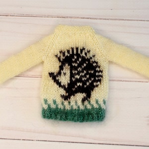 May include: A yellow knitted sweater with a brown hedgehog design and green grass trim.
