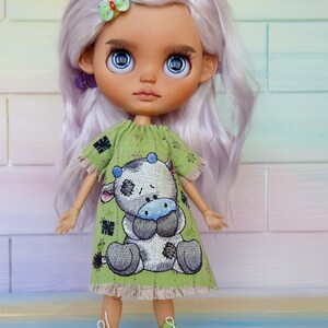 Può includere: Una bambola con lunghi capelli lavanda che indossa un vestito verde con un disegno di una mucca dei cartoni animati. La bambola ha gli occhi blu e indossa scarpe verdi.