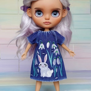 Puede incluir: Una mu&ntilde;eca con el pelo largo y blanco que lleva un vestido de mezclilla azul con flores blancas bordadas y un conejito blanco. La mu&ntilde;eca tiene ojos azules y lleva zapatos blancos.
