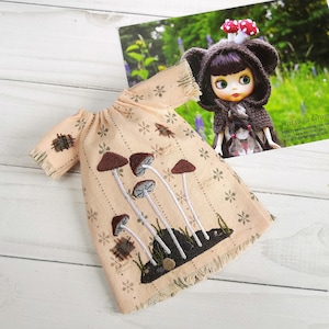 Può includere: Un vestito da bambola color pesca con un ricamo a forma di fungo. L'abito presenta maniche corte e un orlo sfilacciato. Sullo sfondo, una cartolina con una bambola che indossa un abito a tema funghi.