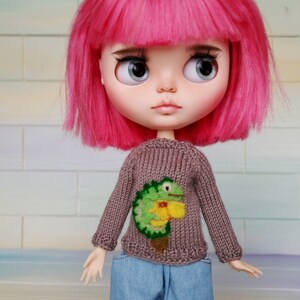 May include: A doll with pink hair wearing a brown sweater with a green and yellow embroidered lizard design. The doll is wearing blue jeans.