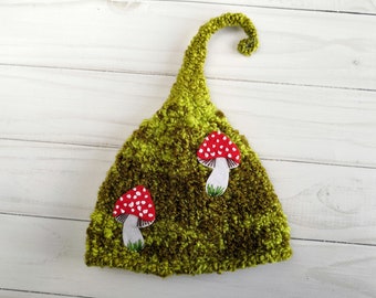 Può includere: Un cappello di lana verde con la cima a punta e un bordo arricciato. Il cappello ha due applicazioni di funghi rossi e bianchi.