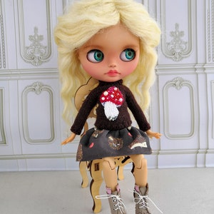 Puede incluir: Una mu&ntilde;eca con cabello rubio que lleva un su&eacute;ter marr&oacute;n con un dise&ntilde;o de hongo, una falda marr&oacute;n con un patr&oacute;n de erizo y botas marrones con detalles rosas.
