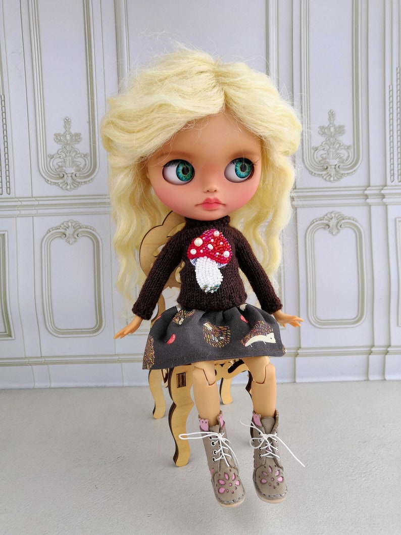 Puede incluir: Una mu&ntilde;eca con cabello rubio que lleva un su&eacute;ter marr&oacute;n con un dise&ntilde;o de hongo, una falda marr&oacute;n con un patr&oacute;n de erizo y botas marrones con detalles rosas.