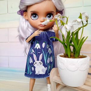 Puede incluir: Una mu&ntilde;eca con el pelo largo y blanco que lleva un vestido de mezclilla azul con un conejo blanco y flores blancas bordadas. La mu&ntilde;eca sostiene una flor blanca y est&aacute; de pie junto a una maceta blanca con plantas verdes.
