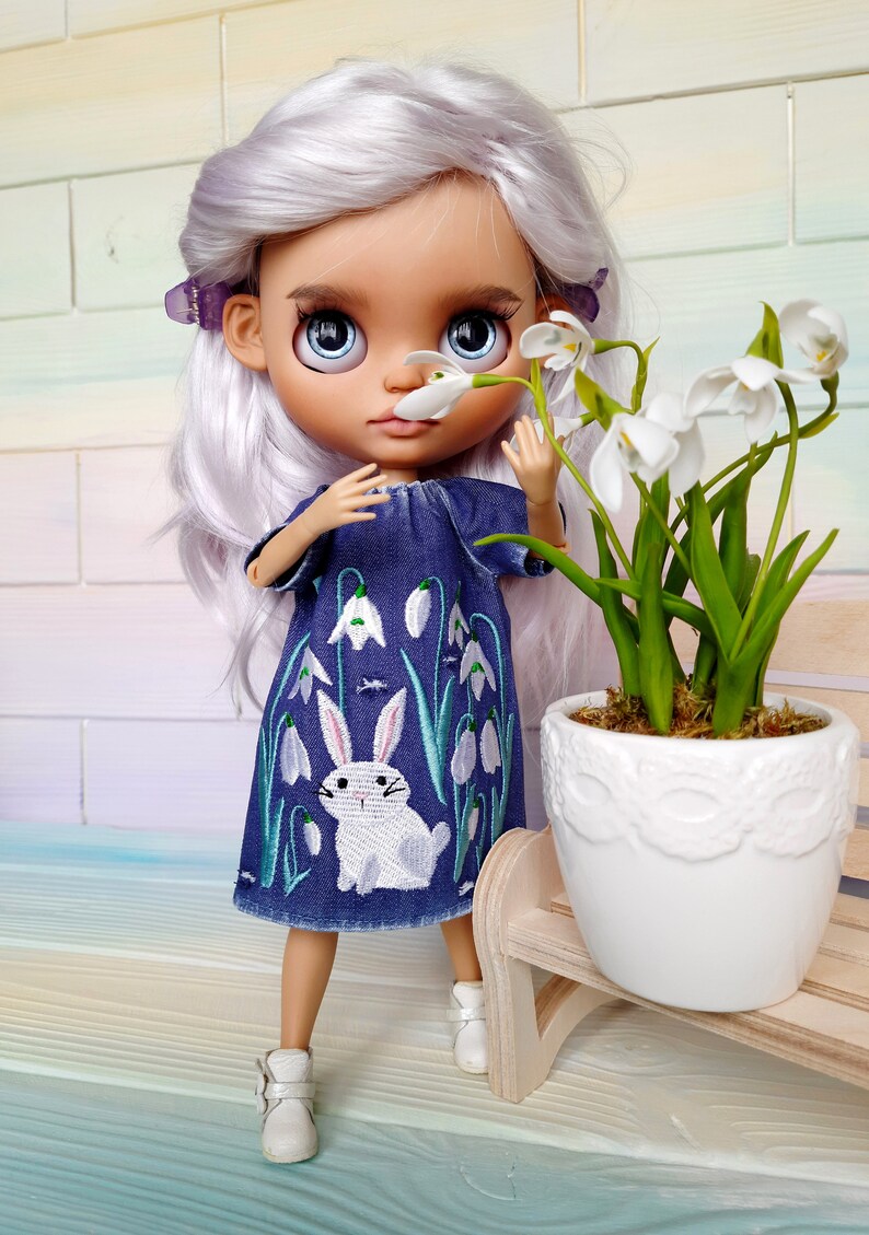 Puede incluir: Una mu&ntilde;eca con el pelo largo y blanco que lleva un vestido de mezclilla azul con un conejo blanco y flores blancas bordadas. La mu&ntilde;eca sostiene una flor blanca y est&aacute; de pie junto a una maceta blanca con plantas verdes.